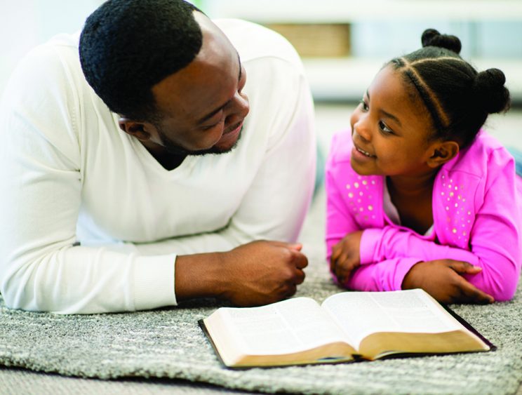 Father and daughter together in the living room reading the bible and discussing religion.