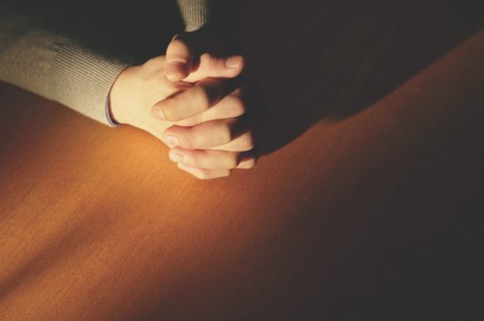 The sun shining on a manâs praying hands on a wooden table, prayer of obedience